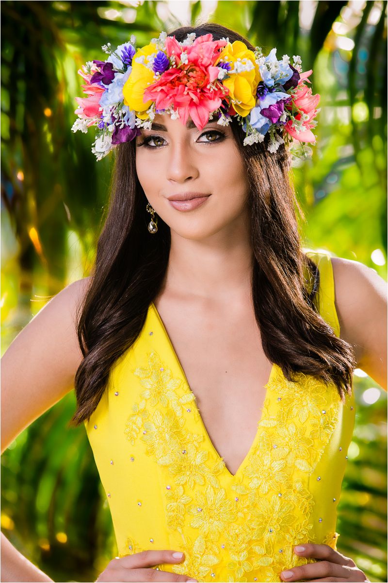 A person wearing a vibrant yellow lace dress and colorful floral crown poses in a tropical garden setting.