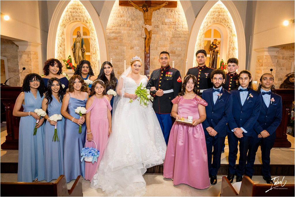 Large wedding party in coordinated formal attire poses in front of decorated church altar.