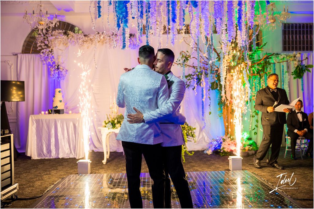 A couple shares a romantic dance beneath colorful lights and glowing floral decor at an elegant evening reception.