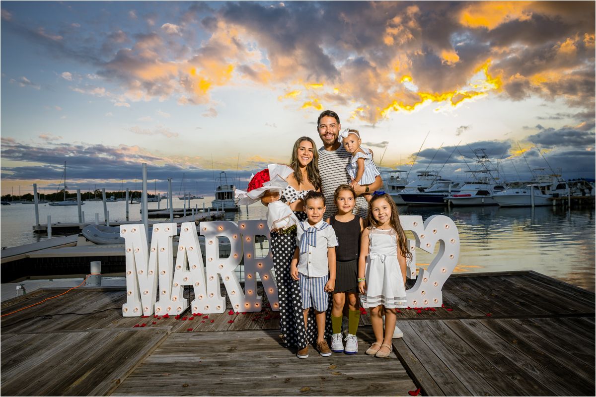 A group poses on a wooden dock at sunset with large 'MARRIED' letters and boats in the marina background.