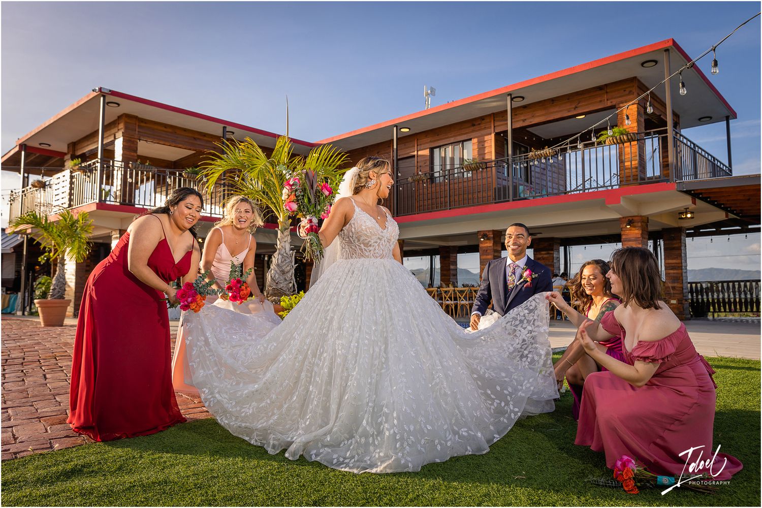 Bridal party in burgundy dresses arranges bride's ballgown on grass outside modern venue.