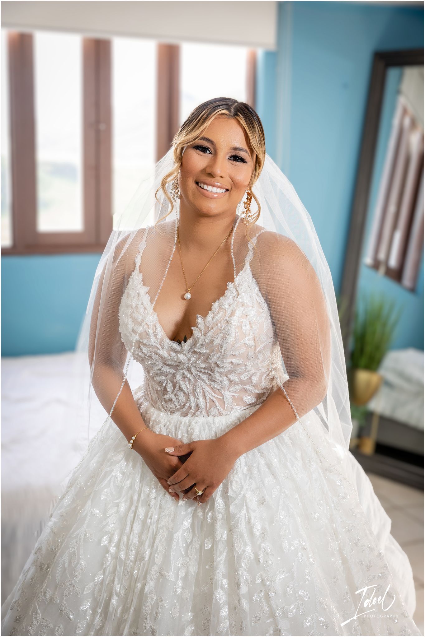 A stunning bride in a white lace wedding gown with delicate veil smiles brightly against a blue wall.