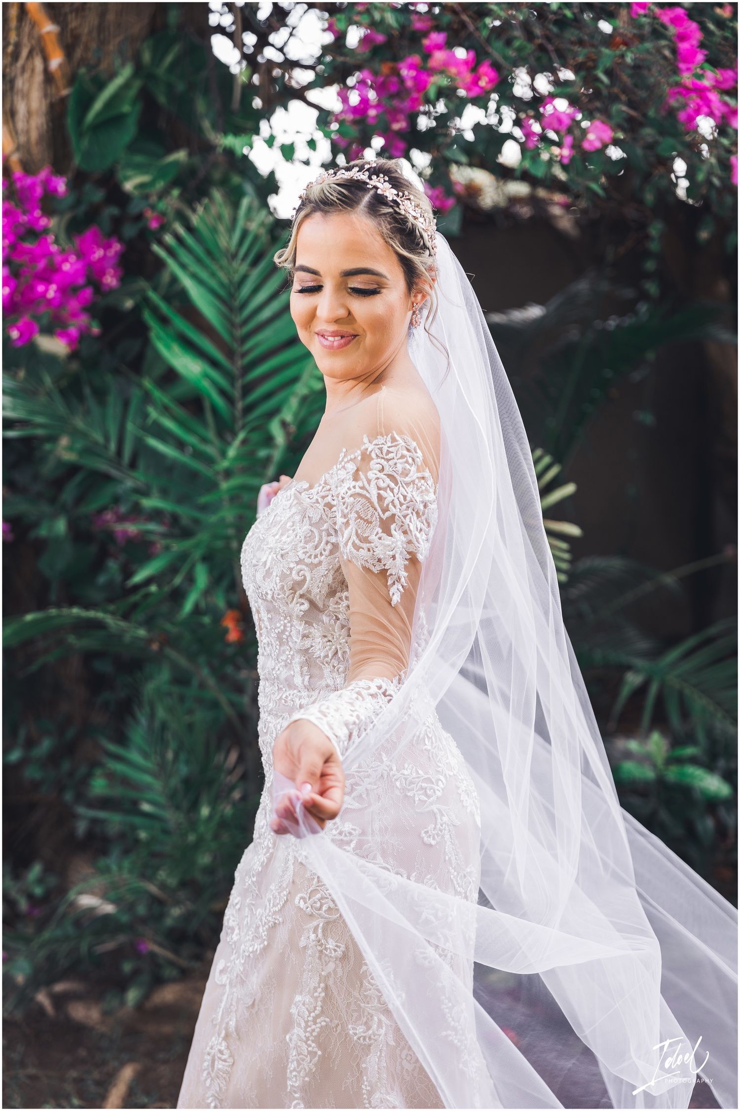 A bride in an ornate lace gown smiles while holding her flowing veil in a garden.