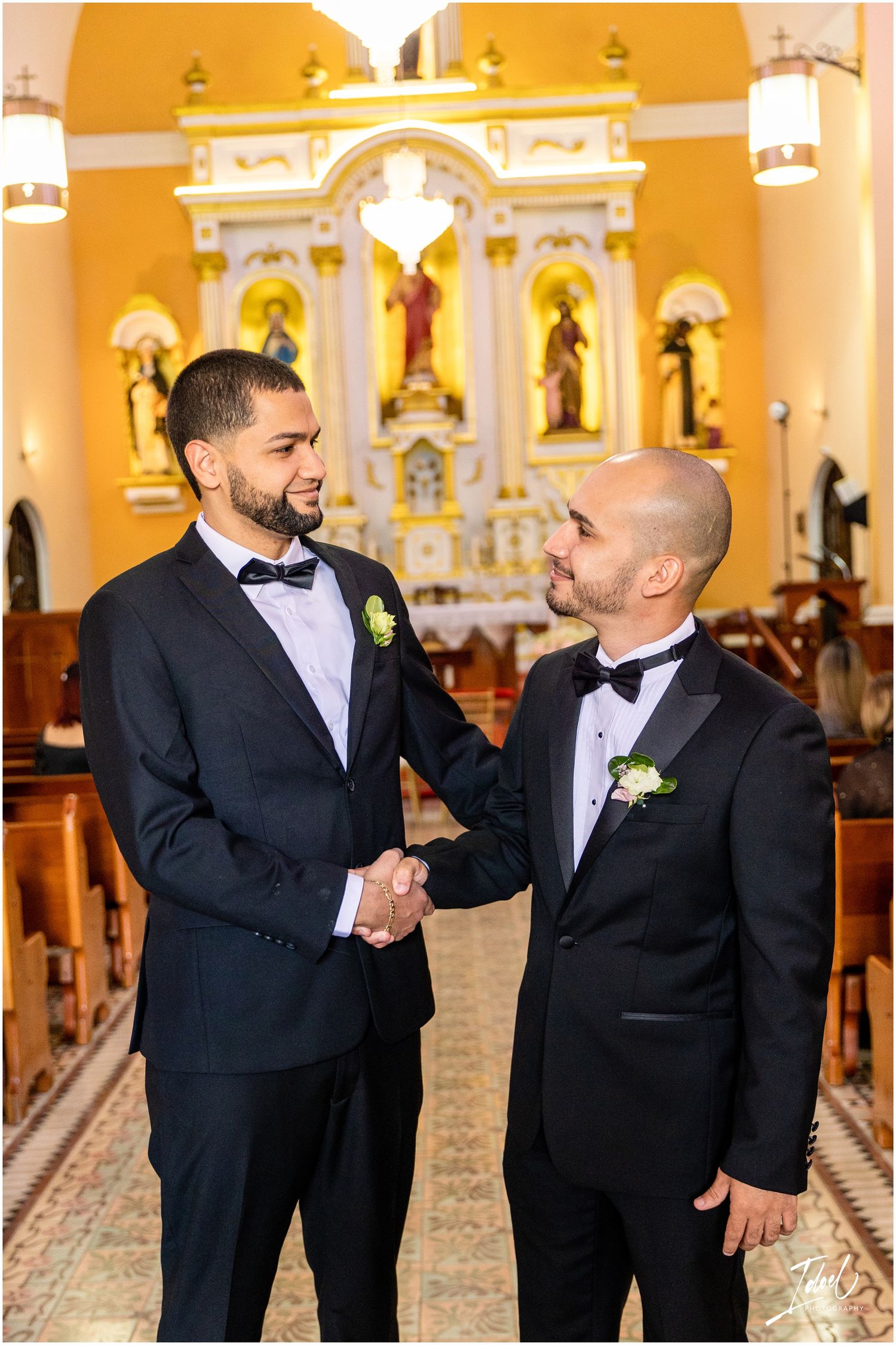 Two formally dressed guests share a handshake and smile inside a church with yellow walls and religious statues.