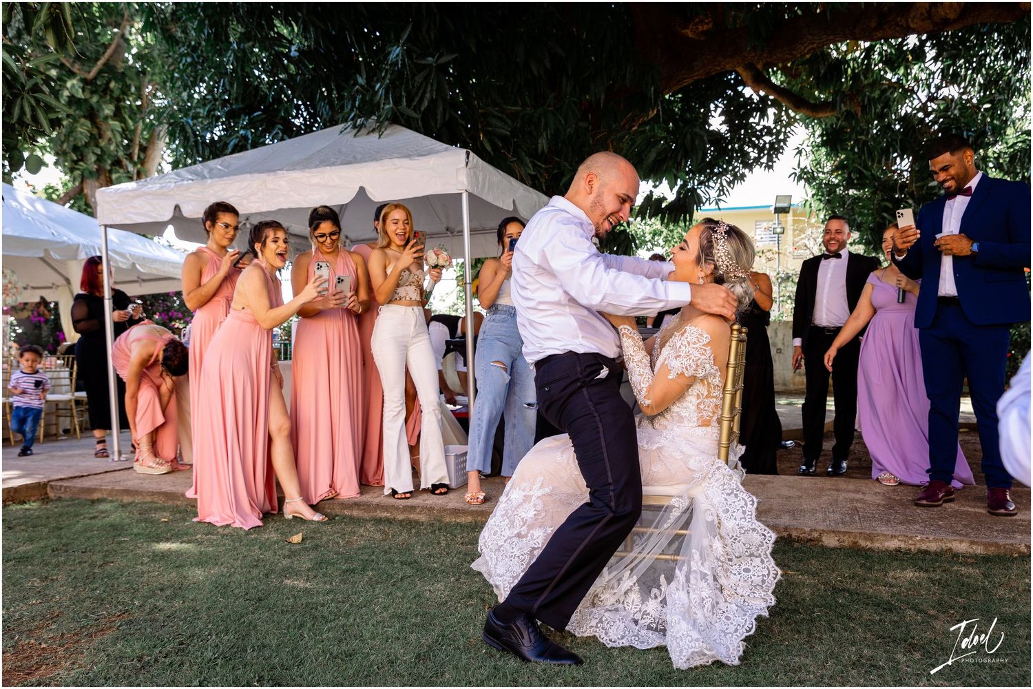 A couple dances outdoors at a wedding reception while wedding party members in pink dresses watch from the sidelines.