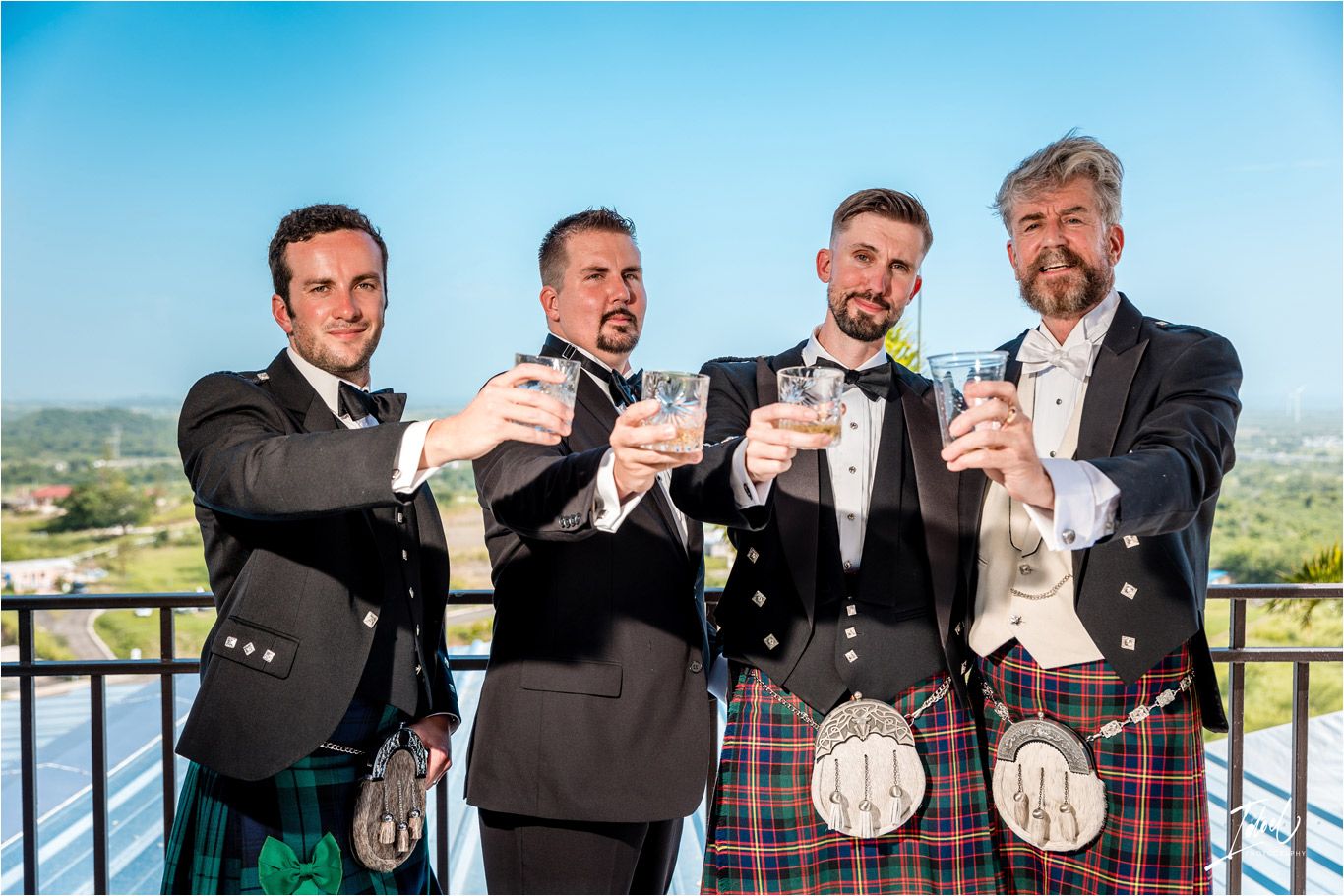 Wedding party members in kilts and formalwear raise glasses in a celebratory toast on a scenic balcony.