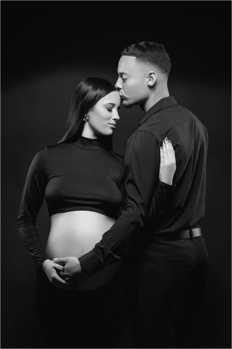 Black and white maternity photo showing intimate moment between couple in dark clothing against dark background.