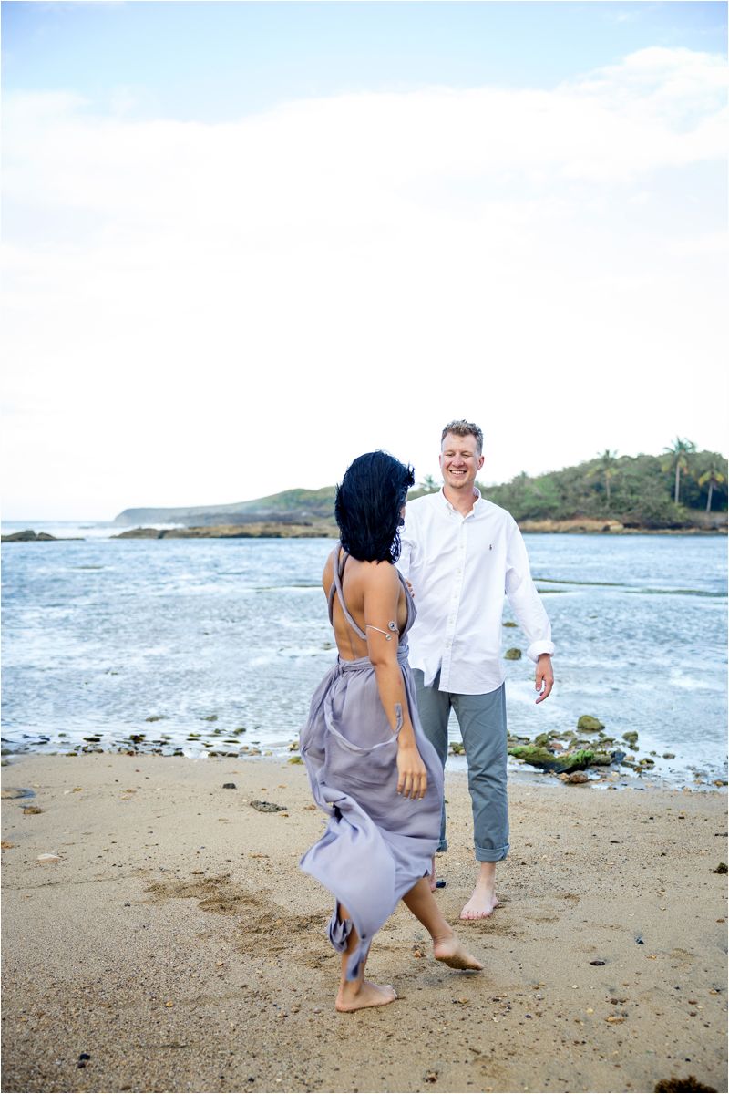 A couple walks playfully along a scenic beach with ocean waves and coastline in the background.