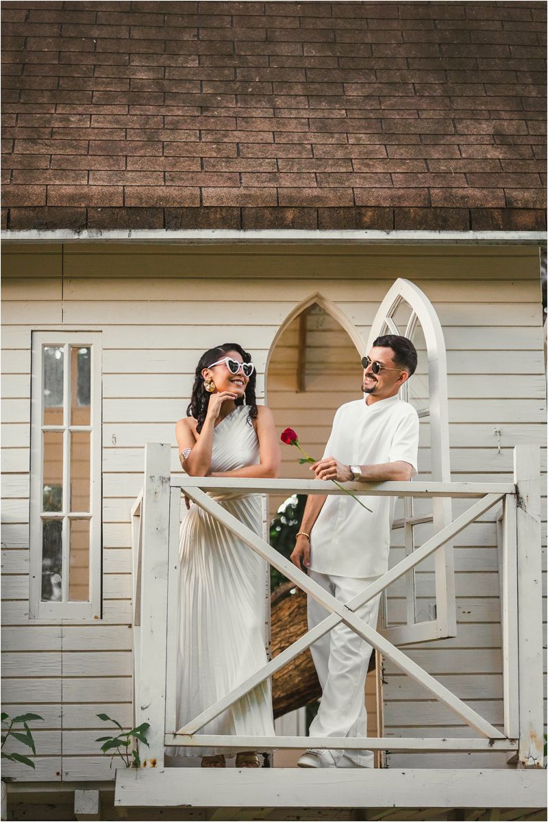 A couple in white attire shares playful moments on a porch with brick and white siding backdrop.