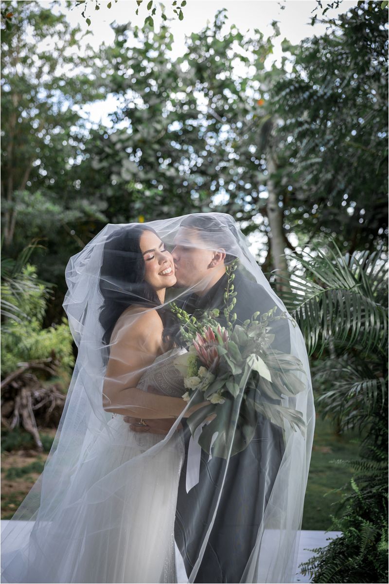 A romantic wedding moment captured in a lush garden setting with flowing white veil and gray attire.