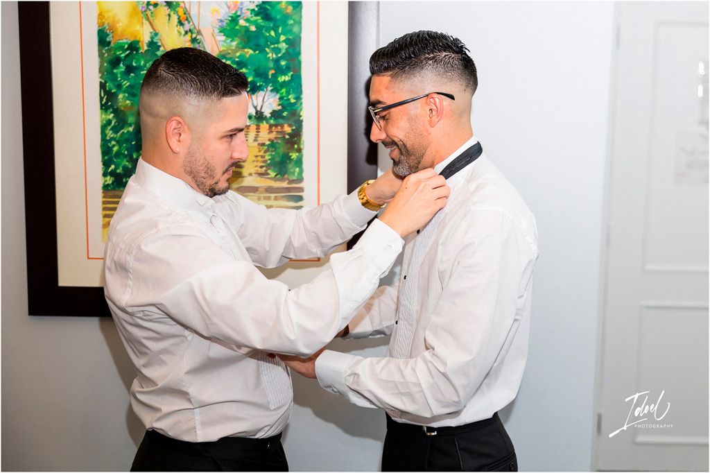 Two people in white dress shirts help each other adjust their bow ties while getting ready for a formal event.