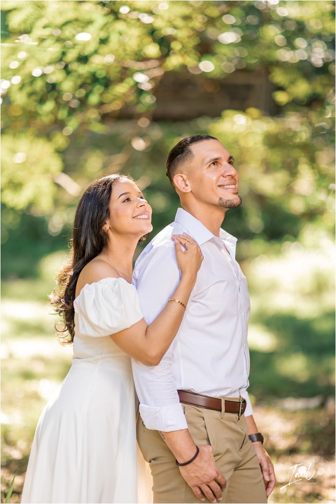 A romantic outdoor engagement photo shows a couple in white and khaki laughing together in dappled sunlight.