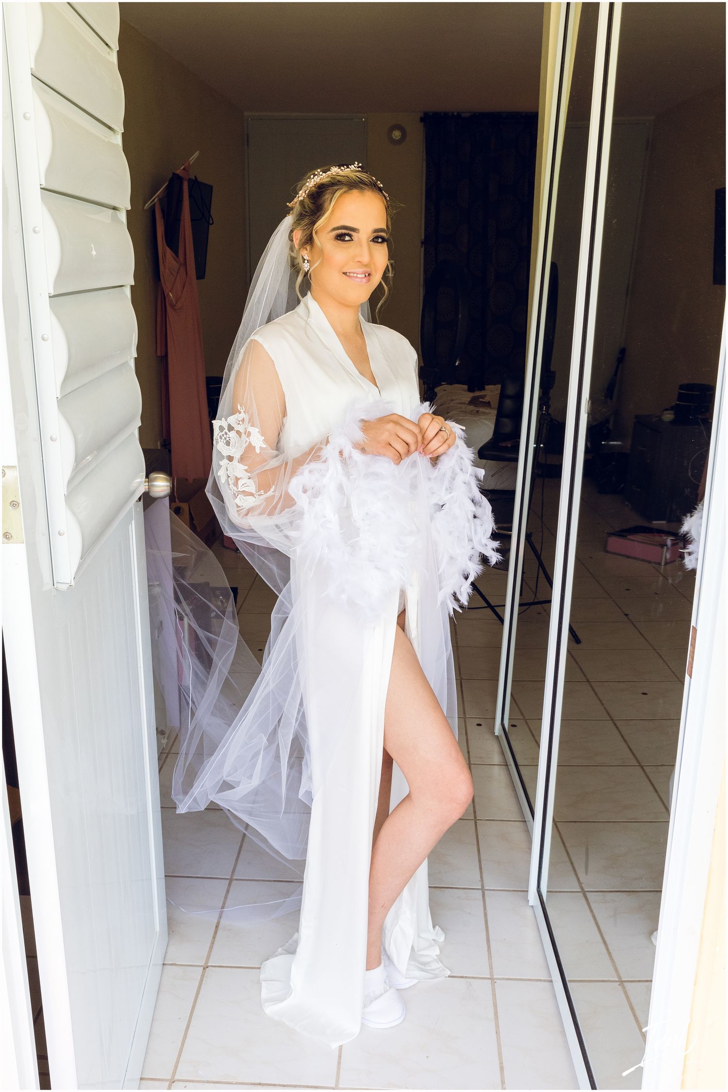 Full-length photo of bride in white silk robe with lace veil standing in doorway.