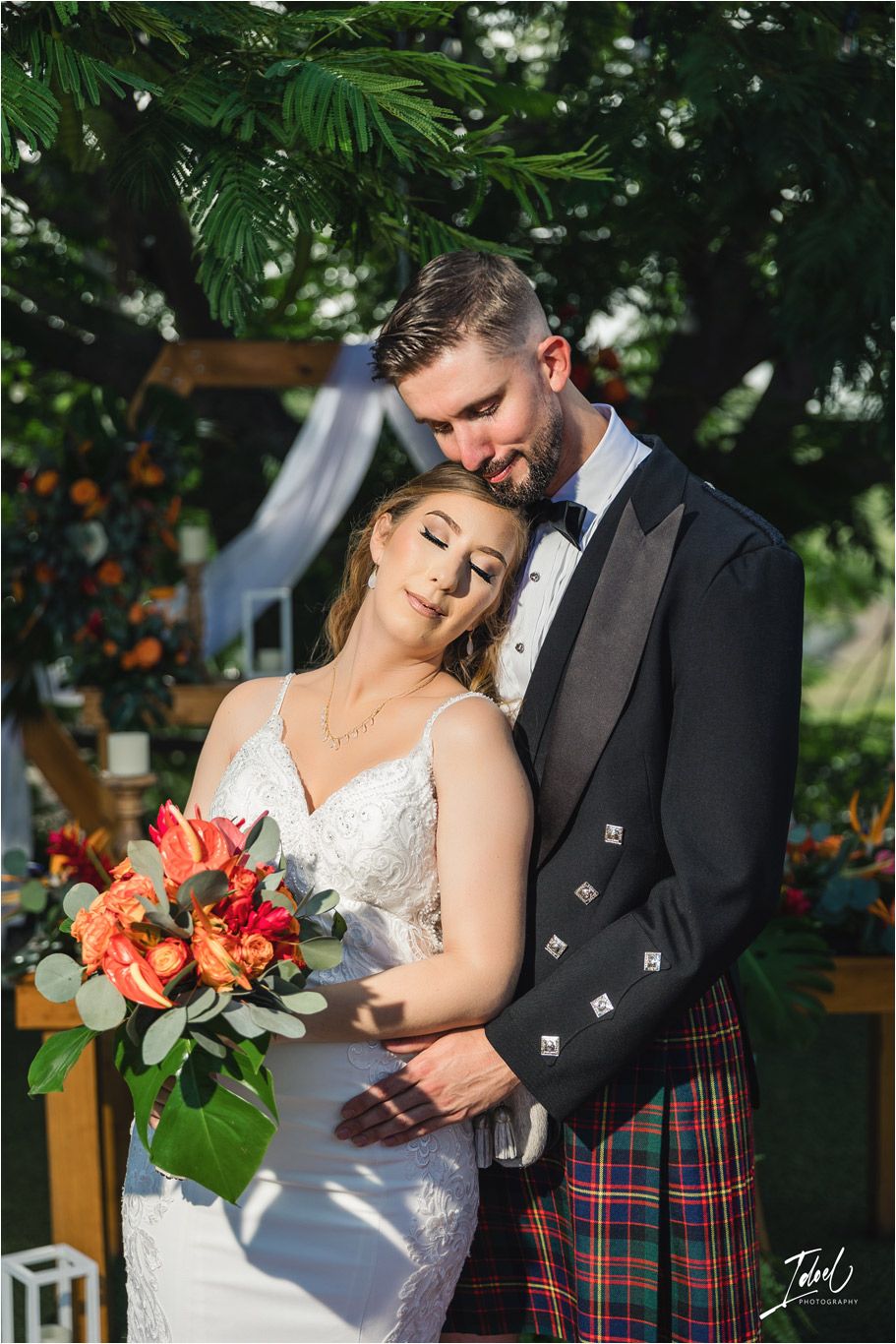 Wedding couple posing under tropical foliage, with bride holding coral bouquet and groom in Scottish kilt attire.