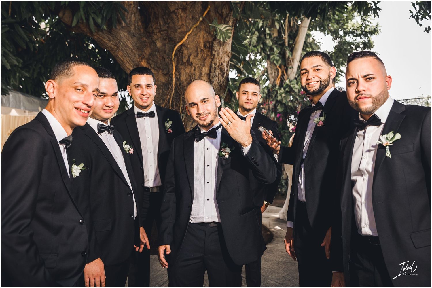 Wedding party in black bow ties and tuxedos shares candid moments during outdoor photo session.
