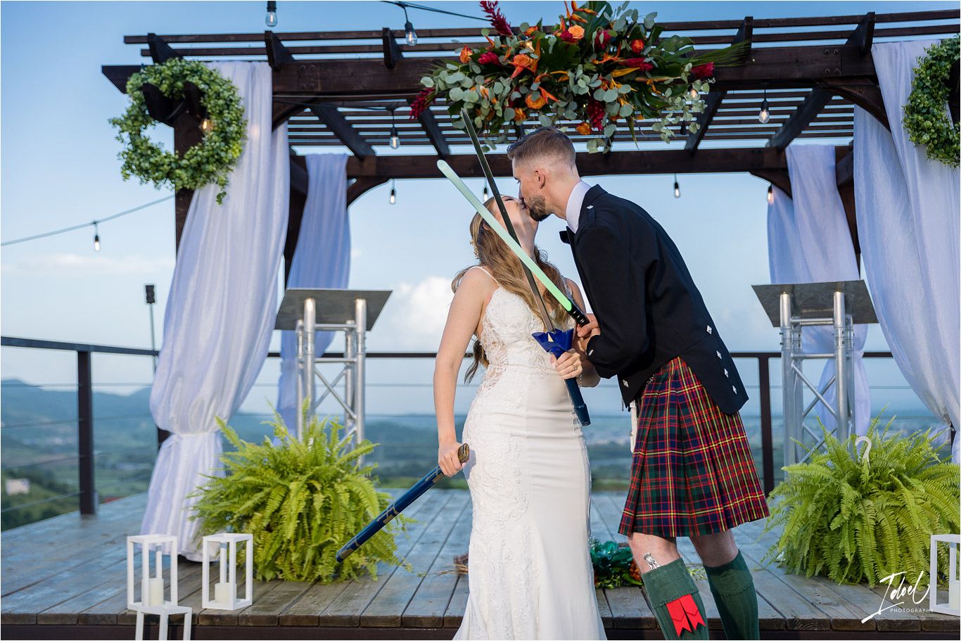 A wedding ceremony under a pergola decorated with floral arrangements and white curtains against a scenic mountain backdrop.