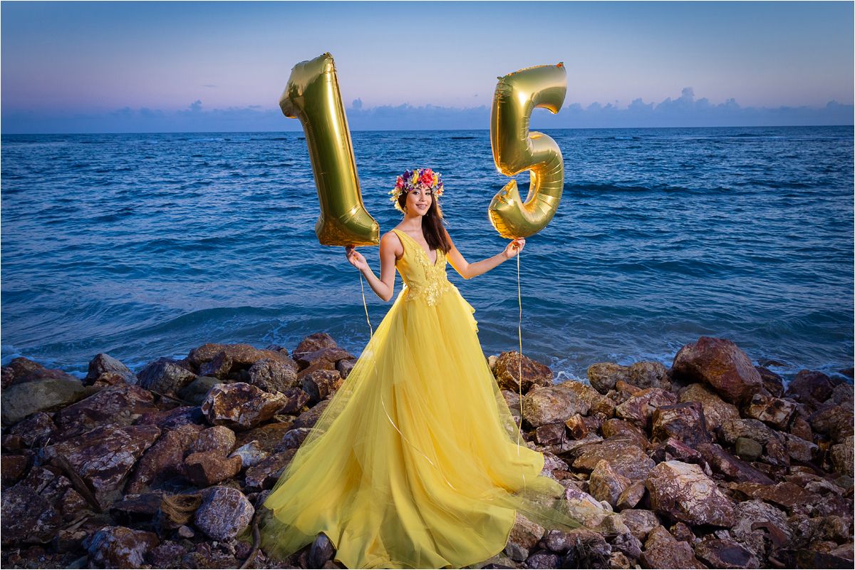 A person in a flowing yellow gown poses gracefully against a rocky beach shoreline at sunset.