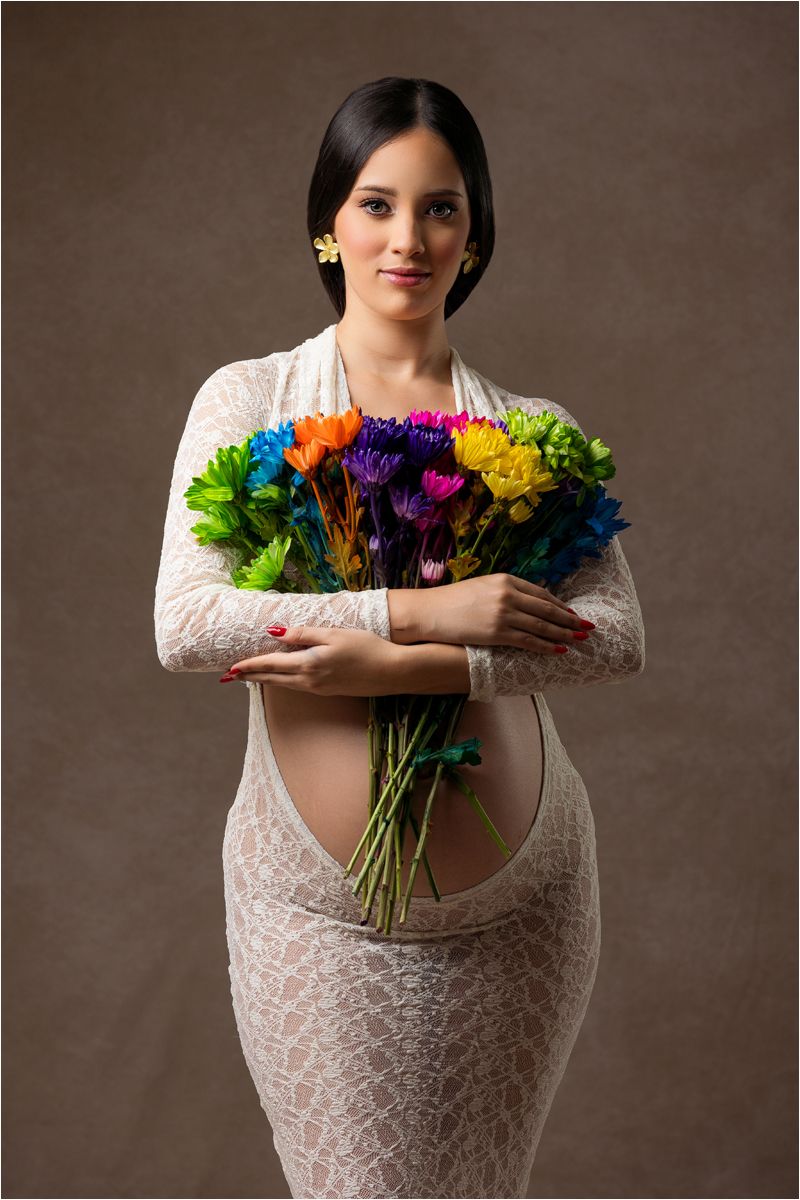 A woman in a white lace dress holds a vibrant bouquet of colorful flowers against a brown backdrop.