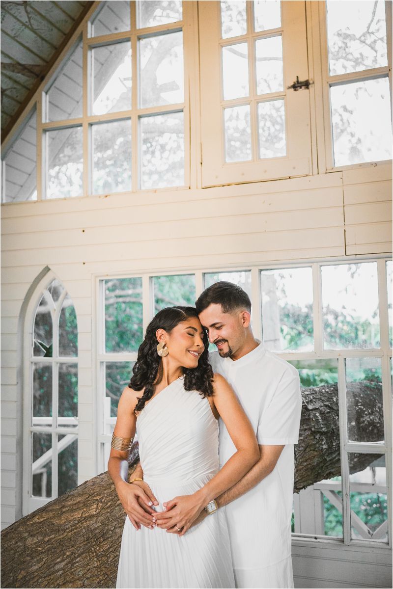 A couple in white outfits share an intimate moment in front of large windows in a bright, rustic chapel setting.