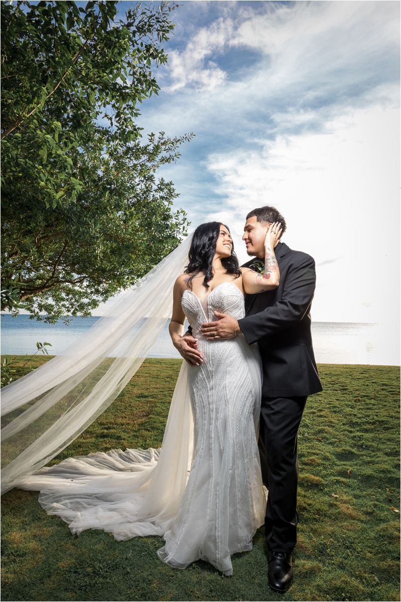 A romantic wedding couple embraces on a grassy hillside as the bride's long veil flows dramatically in the wind.