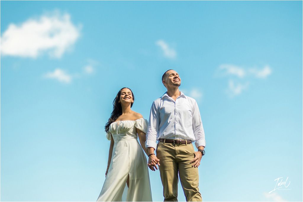 A couple in wedding attire stands holding hands against bright blue sky with wispy clouds.
