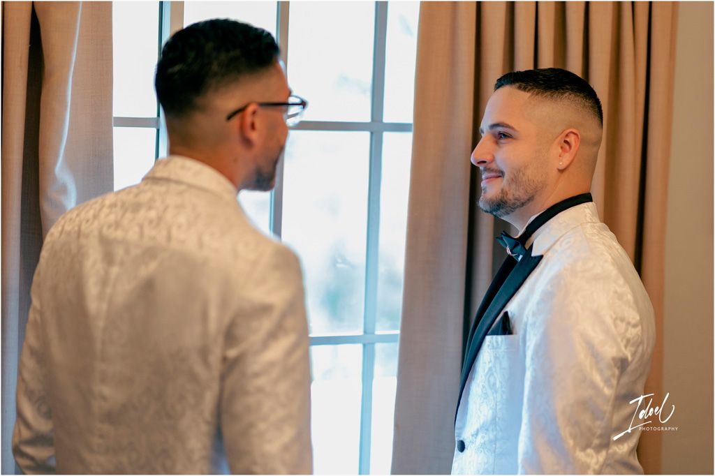 A person in a white suit jacket and bowtie looks at their reflection in a mirror during formal event preparation.