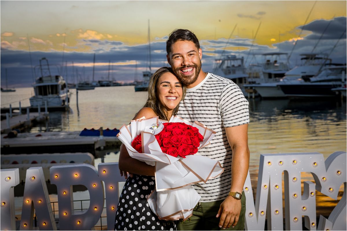 A couple embraces at a marina during sunset while holding a red rose bouquet against a backdrop of docked boats.