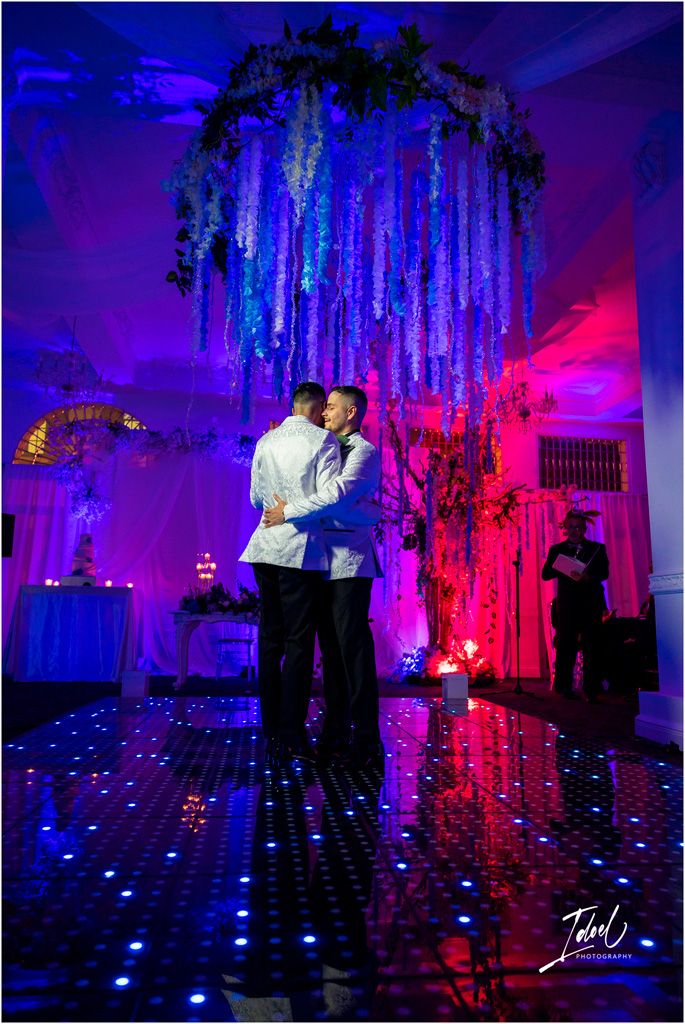 A couple embraces on a reflective dance floor under blue and purple lighting with hanging floral decor in an elegant venue.