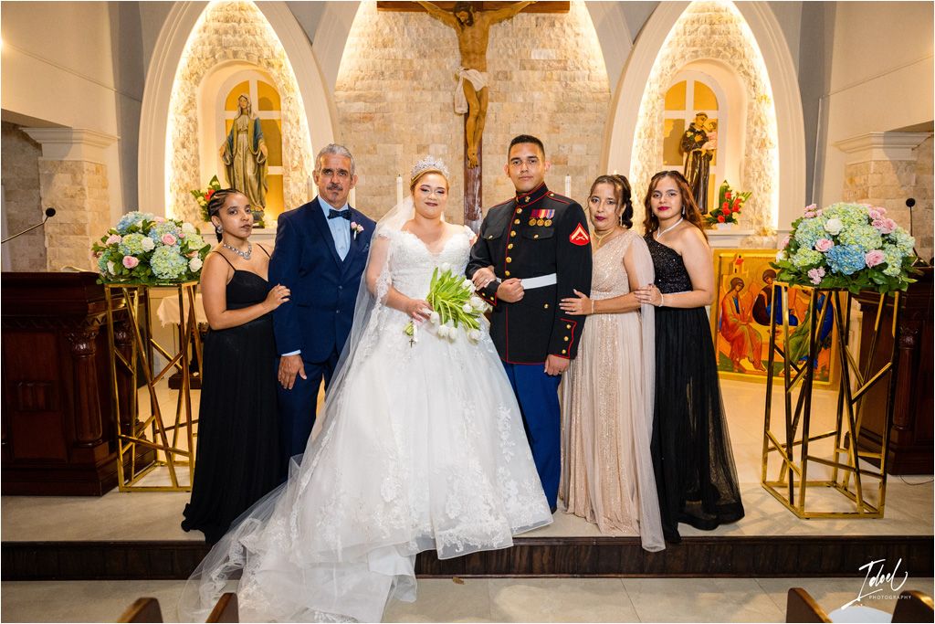 Formal wedding party photo in front of ornate church altar with decorative floral arrangements.
