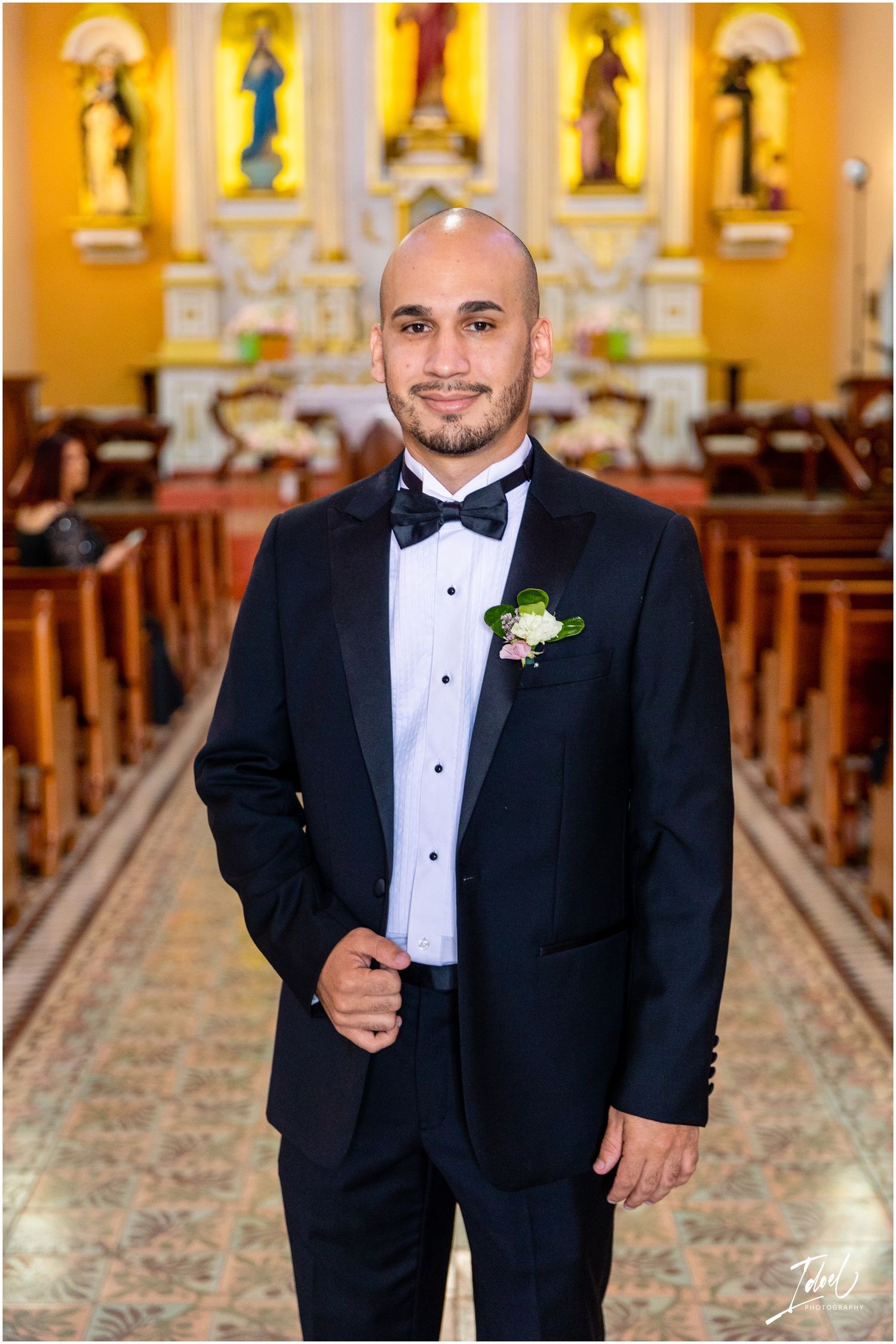 A groom in a black tuxedo and bowtie poses in a Catholic church with ornate statues and yellow walls.