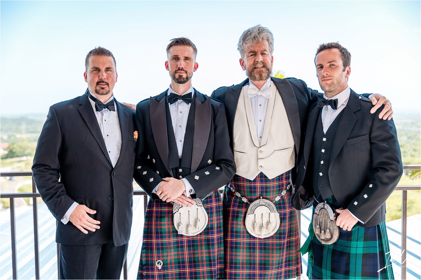 Four groomsmen in formal attire and kilts pose together on a balcony during a wedding celebration.