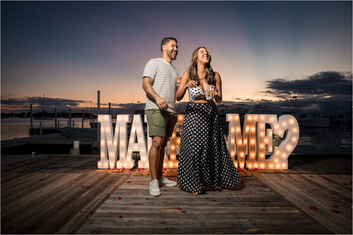 A romantic couple stands on a wooden deck at sunset with illuminated marquee letters spelling 'MARRIED' behind them.