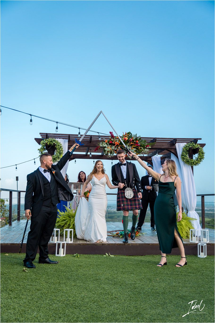 A festive outdoor wedding celebration with string lights and Scottish formal wear against a blue sky.