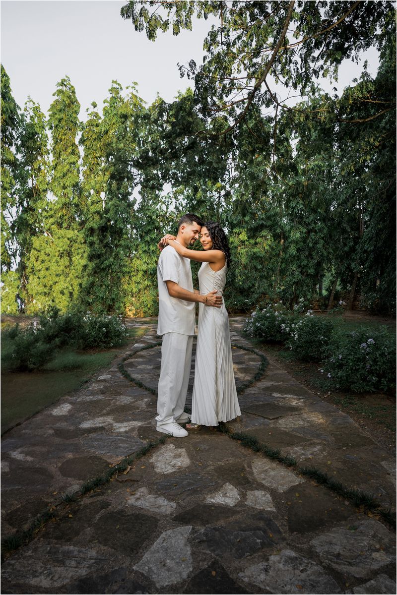 Two people in white dresses embrace in an orchard setting with green trees and a stone pathway.