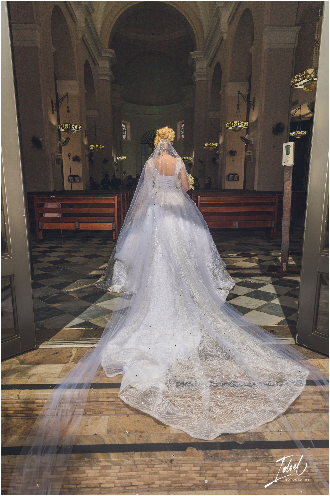 A bride in a lace wedding gown with long train stands in a church with checkered floor.