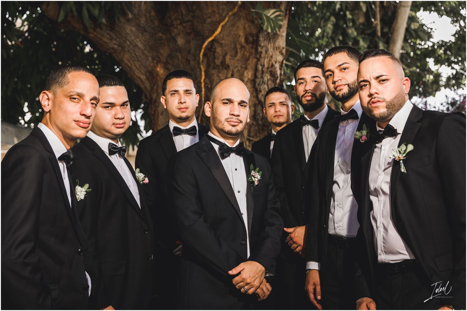 Wedding party of groomsmen in black bow ties and tuxedos gather for outdoor photo.