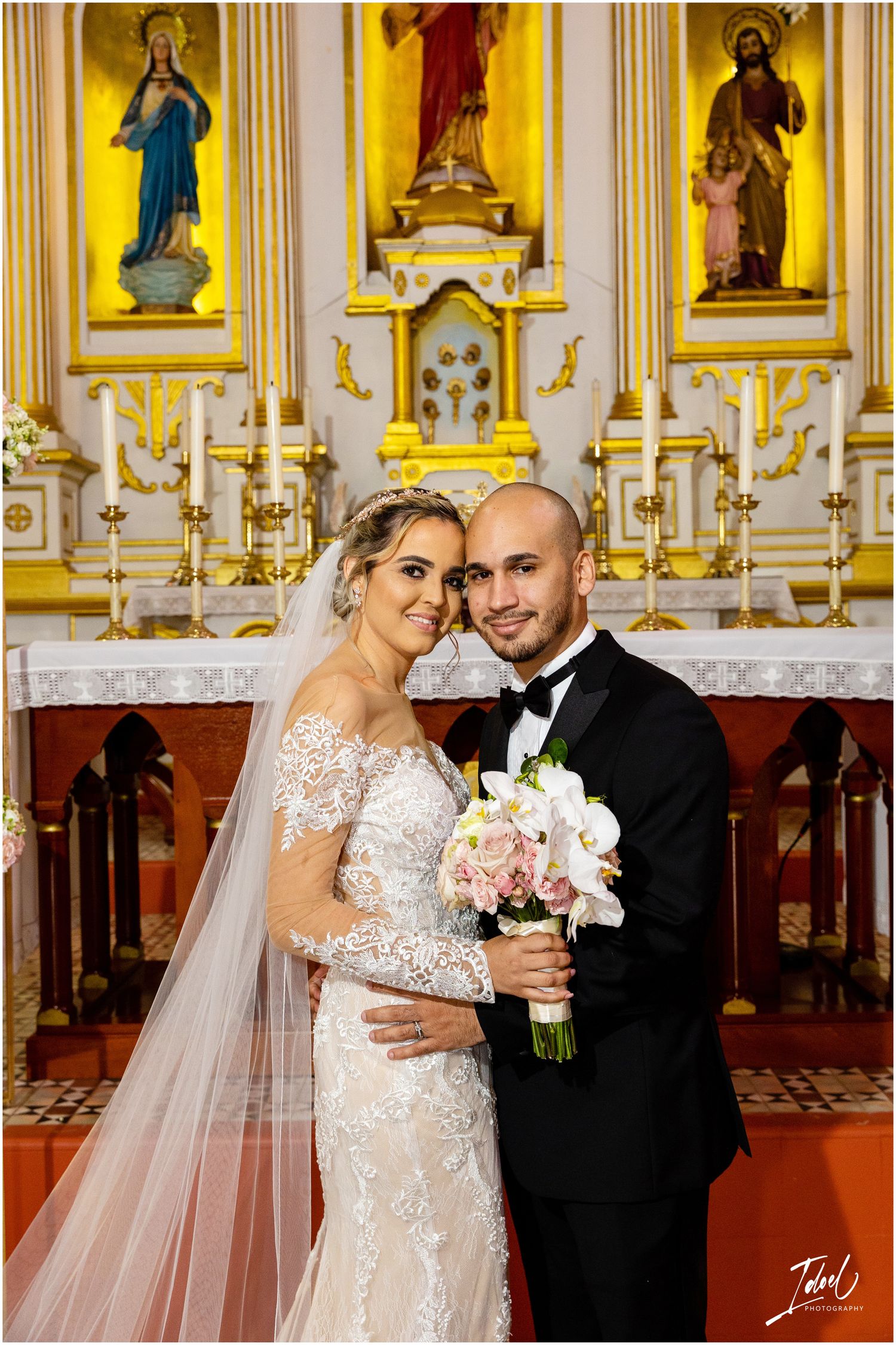 Newlyweds embrace in front of ornate church altar with golden religious paintings.