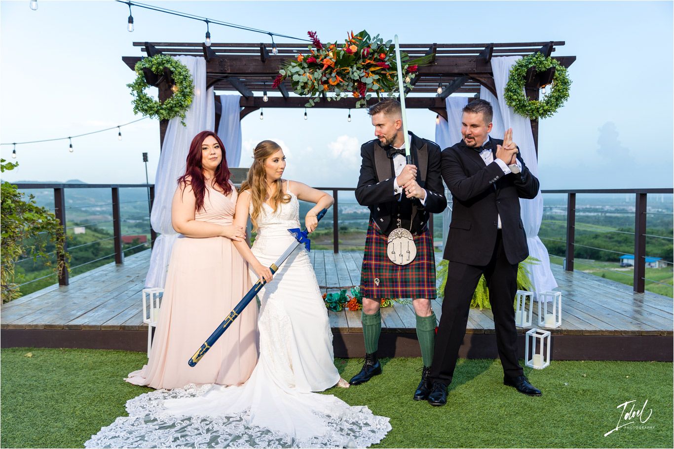 Wedding party poses playfully with swords under decorated outdoor pergola.