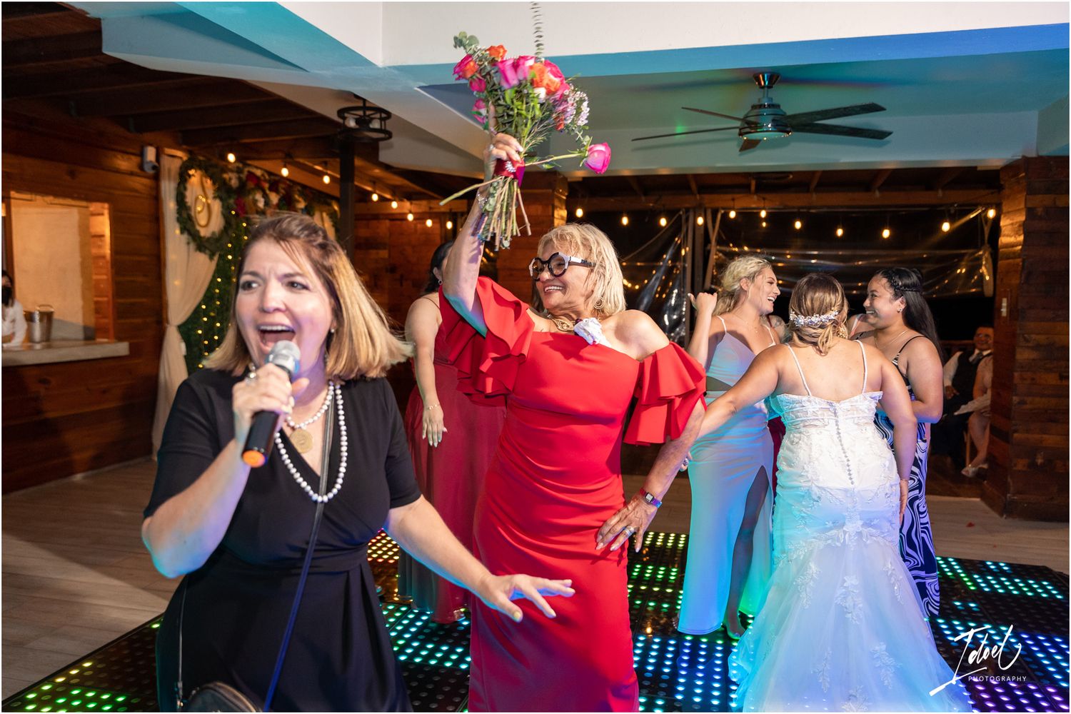 Wedding party celebrating on an illuminated dance floor with raised arms and excited expressions.