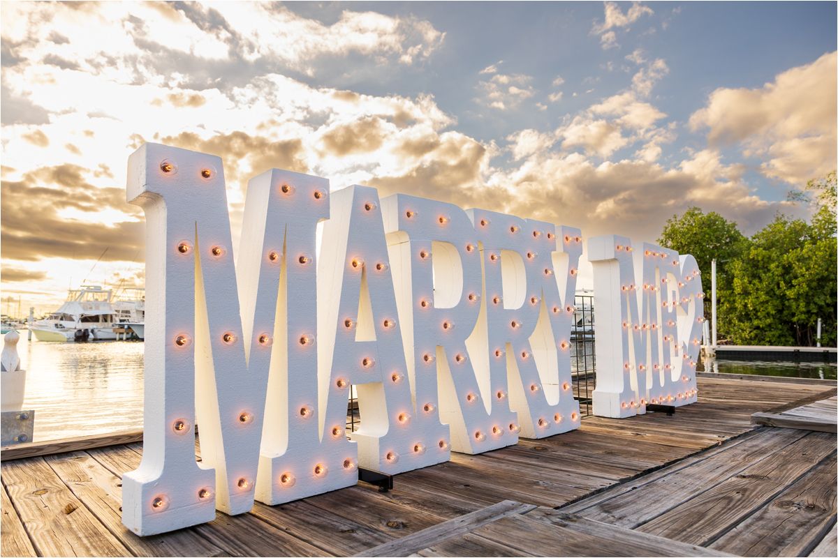 Large illuminated MARRY ME sign with glowing bulbs stands on a wooden deck at sunset with dramatic clouds in the sky.