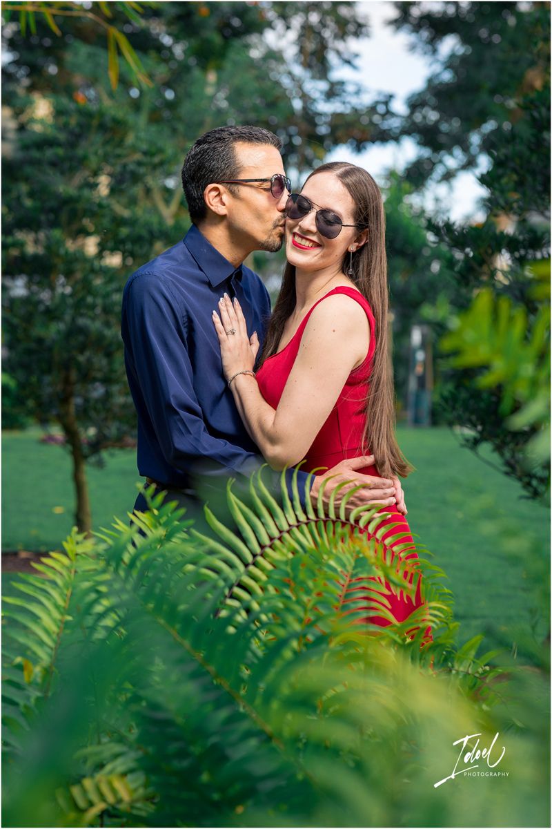 Couple sharing a romantic kiss in a lush green garden, woman in red dress with sunglasses smiling.