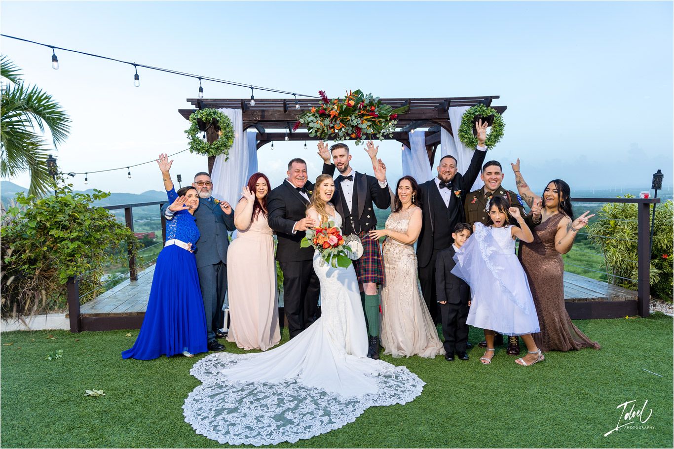 Large wedding group gathers for formal photo under decorated outdoor pergola with mountain views.