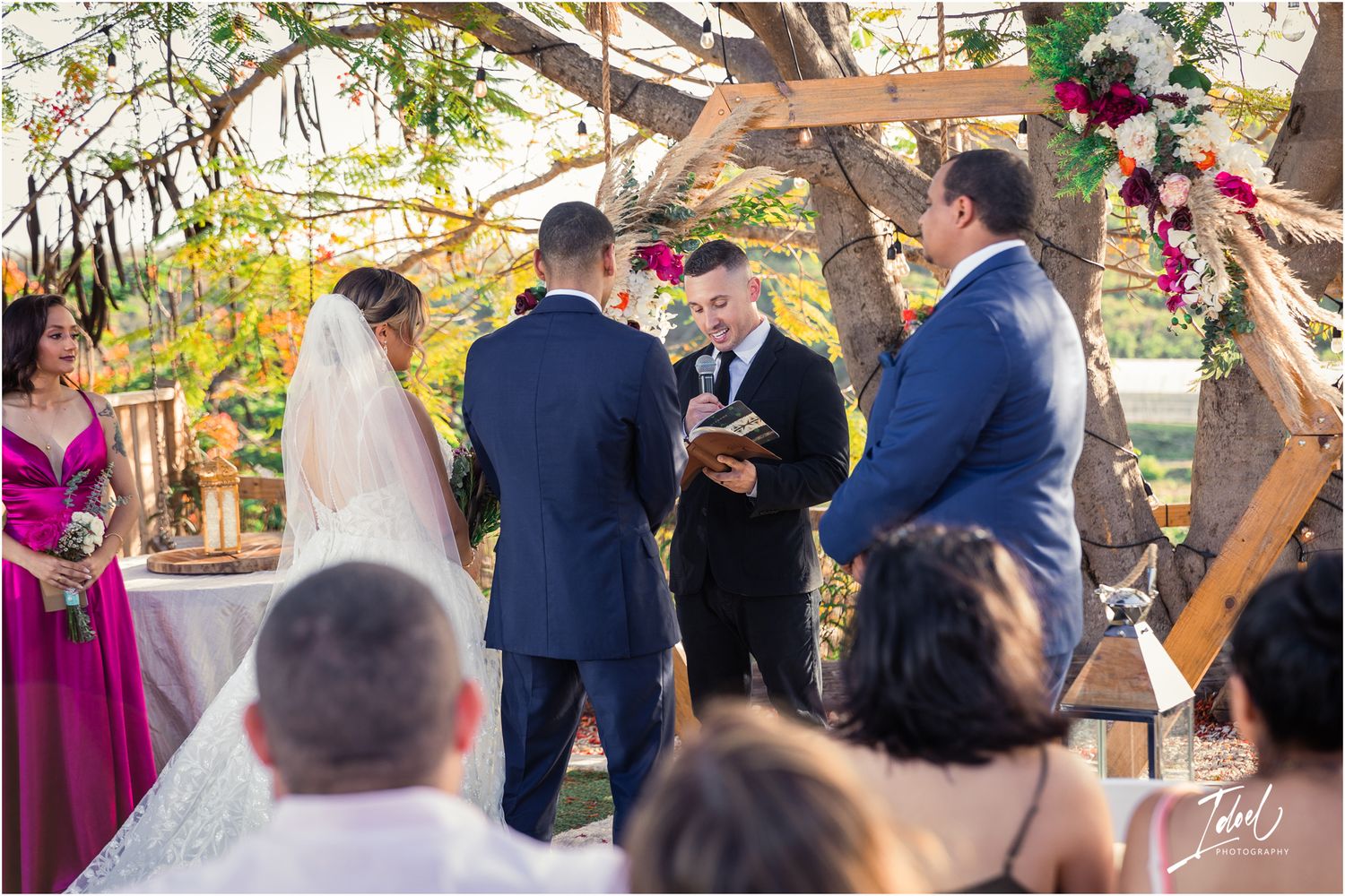 An outdoor wedding ceremony takes place under a rustic wooden arch with pink floral decorations.
