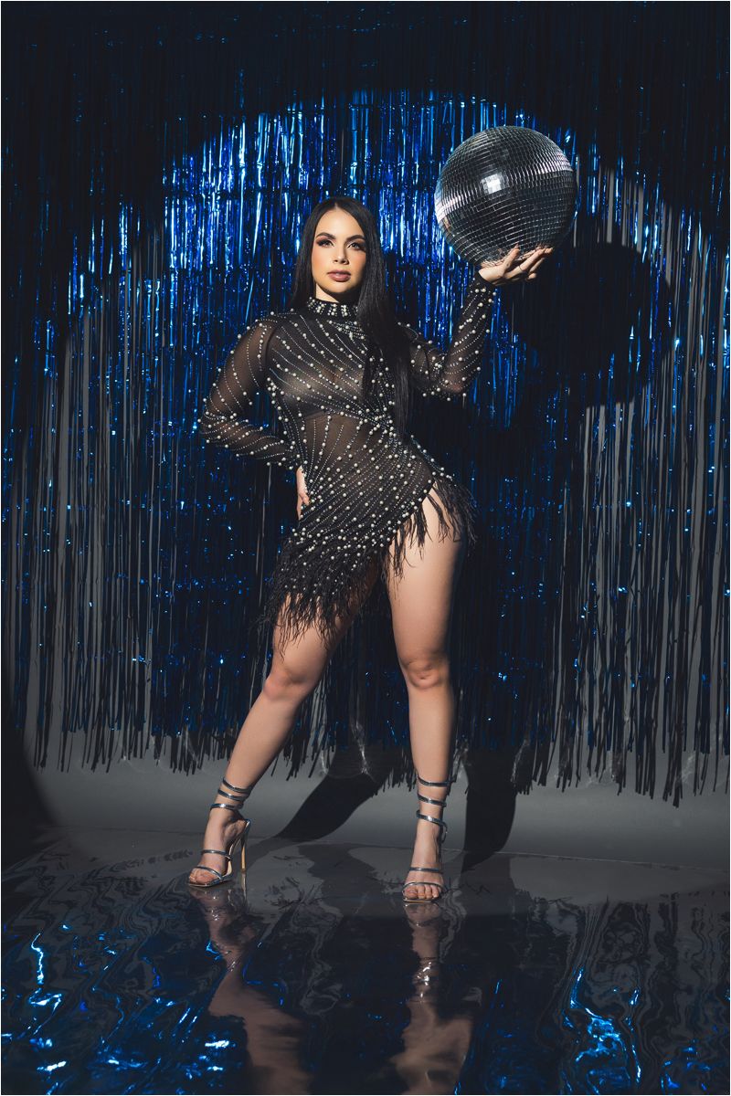 A person in a sparkly black dress poses against a dramatic blue tinsel backdrop in a fashion photoshoot series.