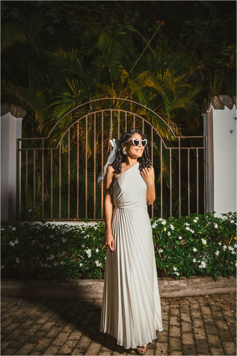Elegant pleated maxi dress photographed against ornate iron gates and tropical foliage in evening light.