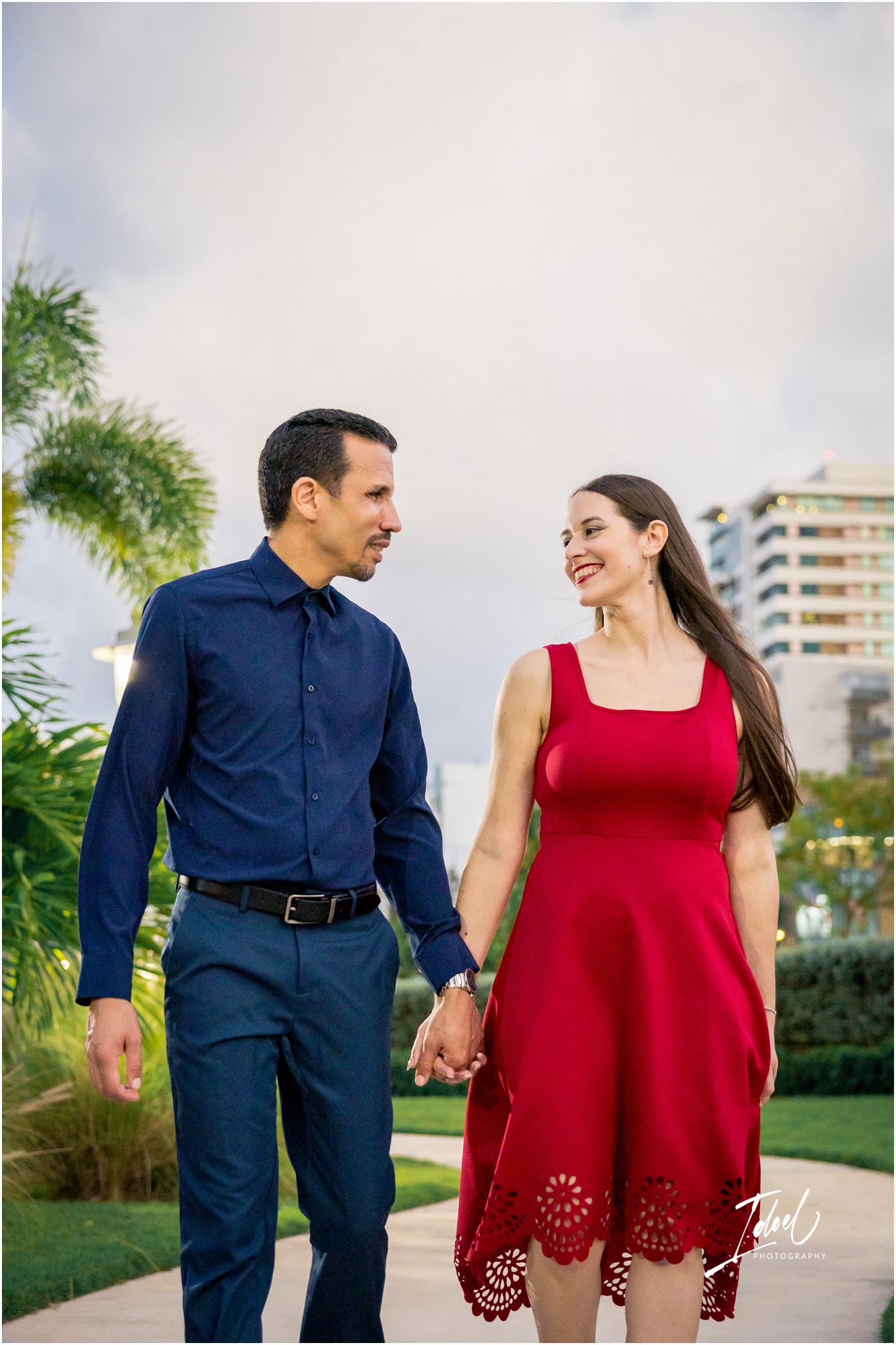 Couple holding hands walking outdoors, man in navy shirt and woman in red dress, smiling at each other near tropical greenery.