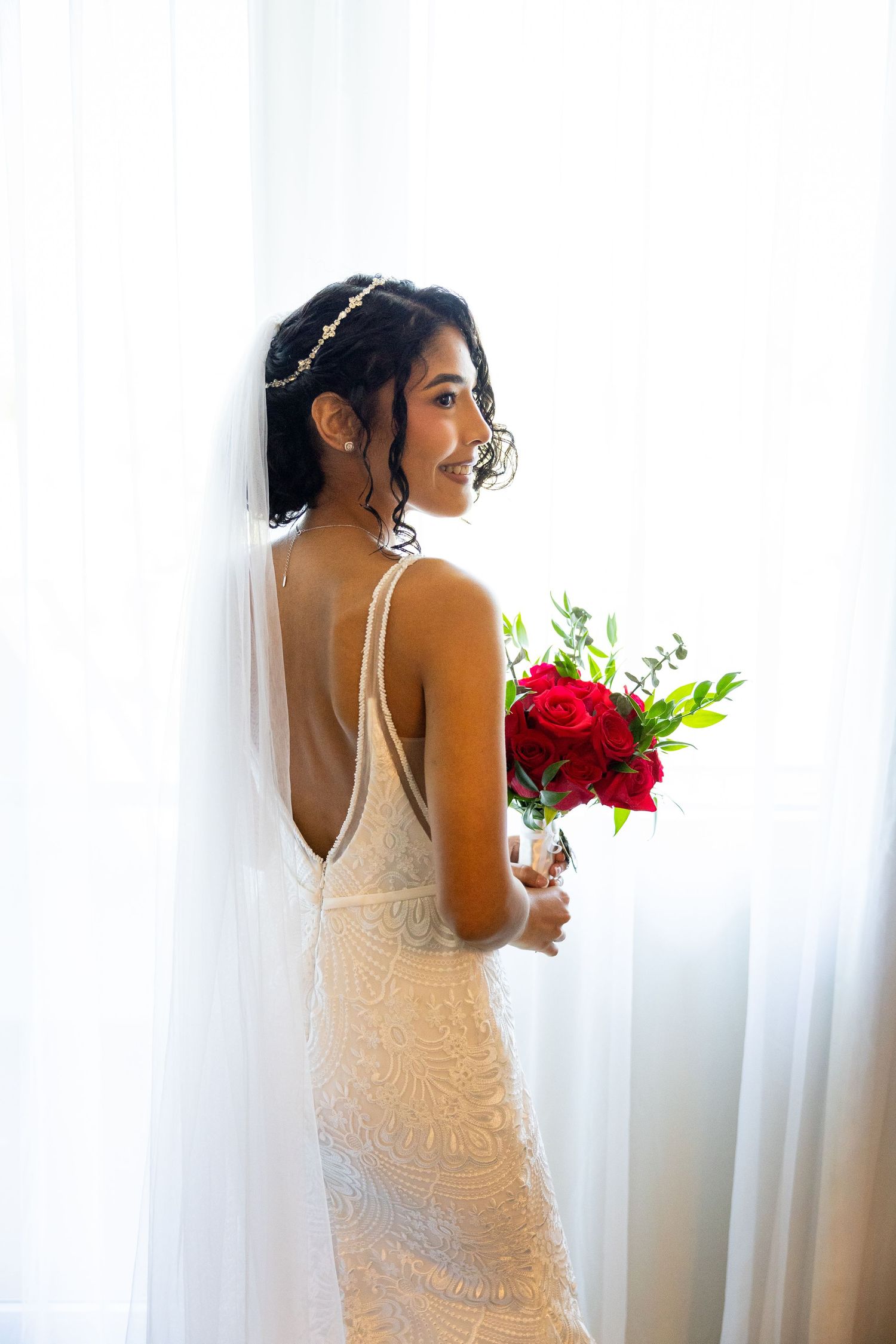 Elegant lace wedding dress with delicate straps shown from behind against white curtains.
