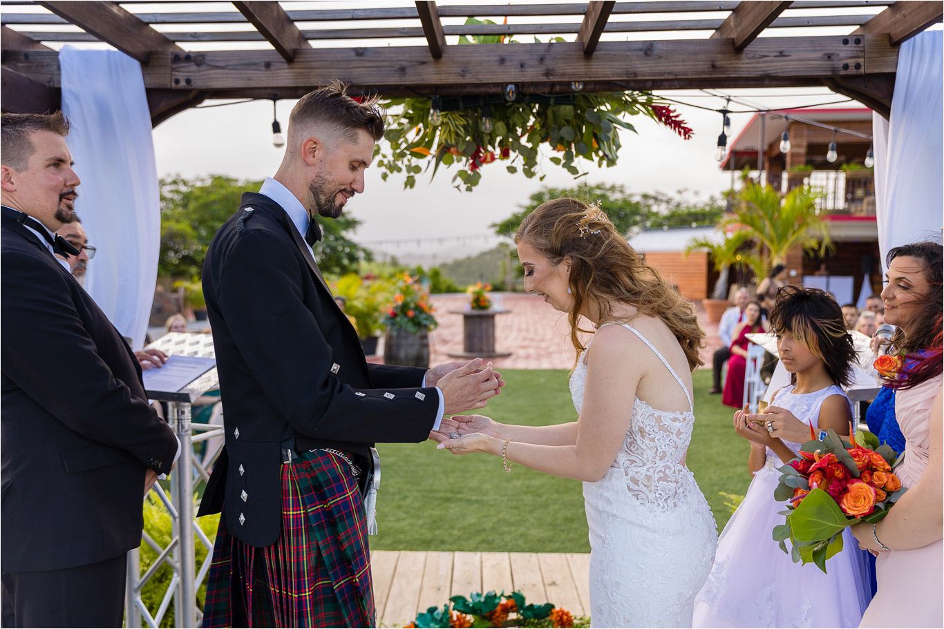 Outdoor wedding ceremony under a pergola with guests watching the proceedings.
