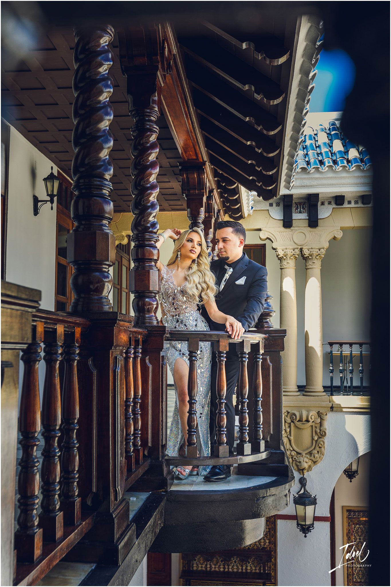 Couple stands on vintage wooden balcony with carved pillars and architectural details.