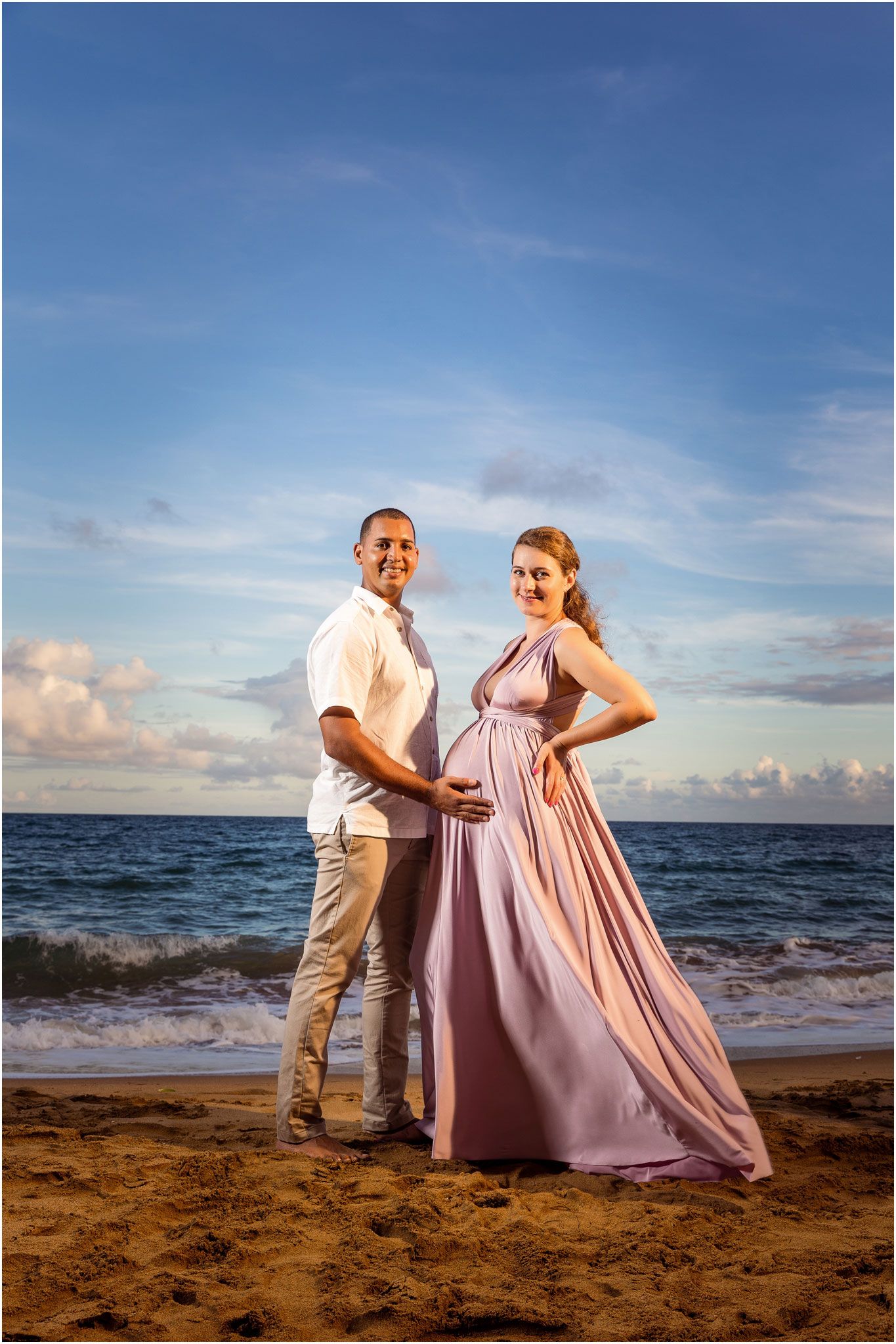 Maternity beach portrait with couple in pink gown and white shirt against ocean backdrop.